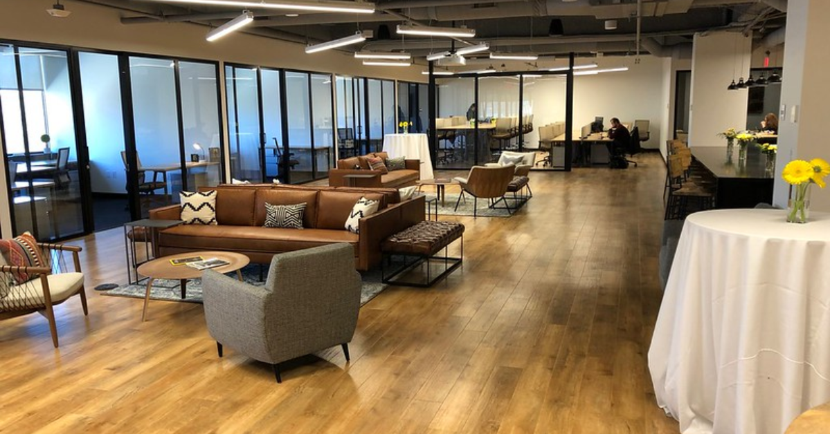 Modern office lounge with brown leather couches, chairs, and tables. Glass-walled meeting rooms line the left side. The wooden floor complements the contemporary decor, creating a warm coworking community vibe for those seeking innovative office space solutions.