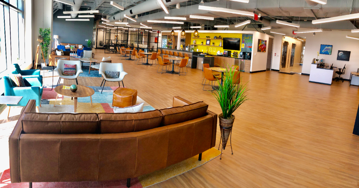 Open office space with brown sofas, colorful rugs, and a yellow wall with a counter sets the tone for this vibrant coworking space. Various chairs and tables are scattered throughout, complemented by plants and artwork for decor.