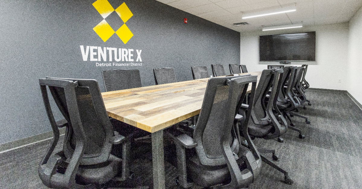 A conference room within a coworking space features a long wooden table, black mesh chairs, and a wall-mounted TV, with the Venture X logo proudly displayed on the wall.
