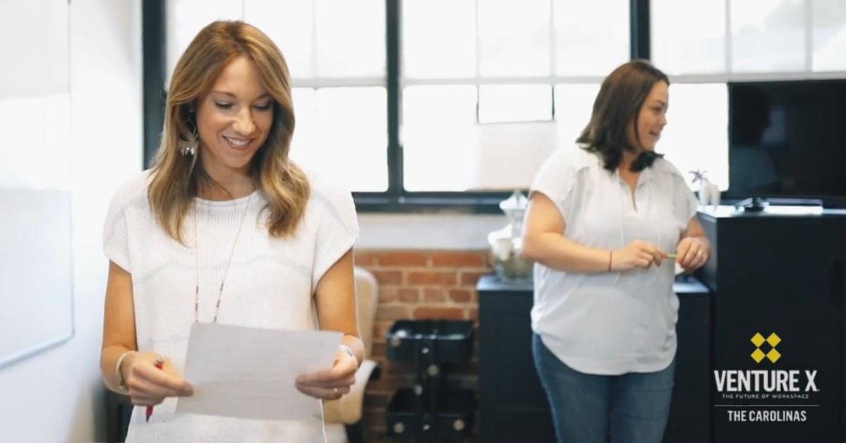 Two women in a bright office; one reads a paper while the other stands near a counter, exemplifying the collaborative spirit of private office space. The Venture X The Carolinas logo graces the corner, embodying modern flexibility and innovation in office space solutions.