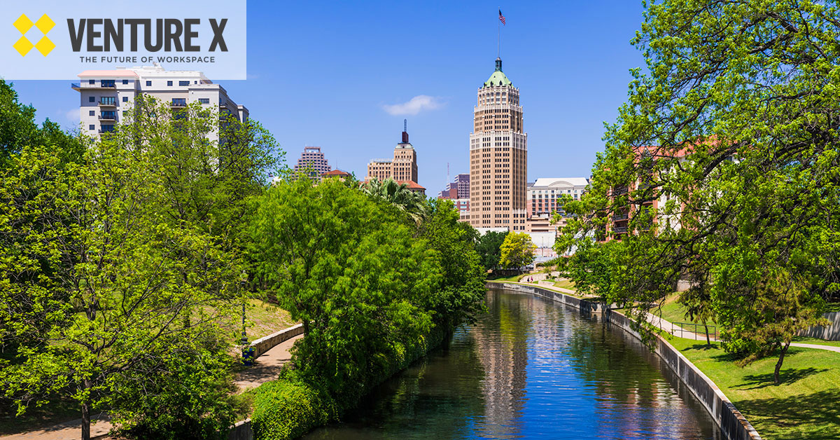Cityscape with a river, lush trees, and tall buildings under a blue sky. Venture X logo in the top left corner, highlighting their flexible workspace solutions amidst this vibrant urban setting.