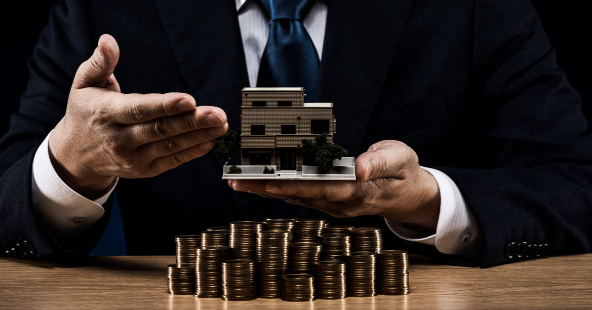 A person in a suit holds a small model house above a stack of coins on the table, contemplating the value of private office space.