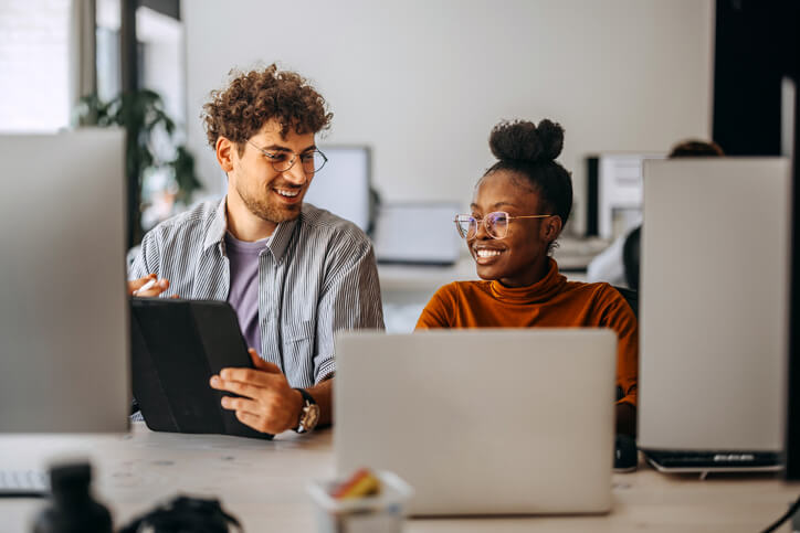 Two colleagues sit at a desk in an office, engaging in conversation. One holds a tablet, and both have open laptops in front of them. They appear to be collaborating on a project. future of work