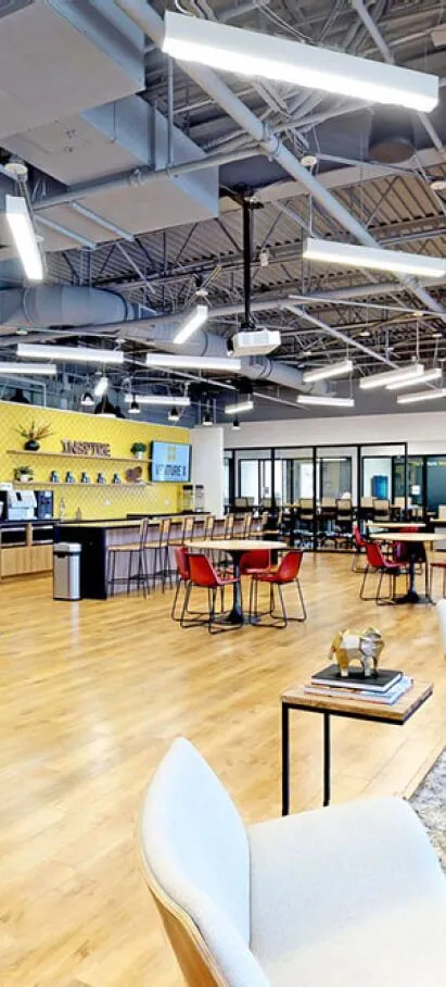 Spacious office lounge with wooden flooring, modern lighting, tables, and red chairs. A vibrant coworking community thrives here with a coffee station against a yellow wall and glass-walled meeting rooms offering flexible workspace options for dynamic professionals.
