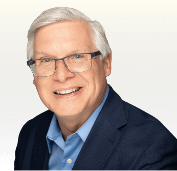 A smiling man with gray hair and glasses wearing a blue shirt and dark blazer against a gradient background.