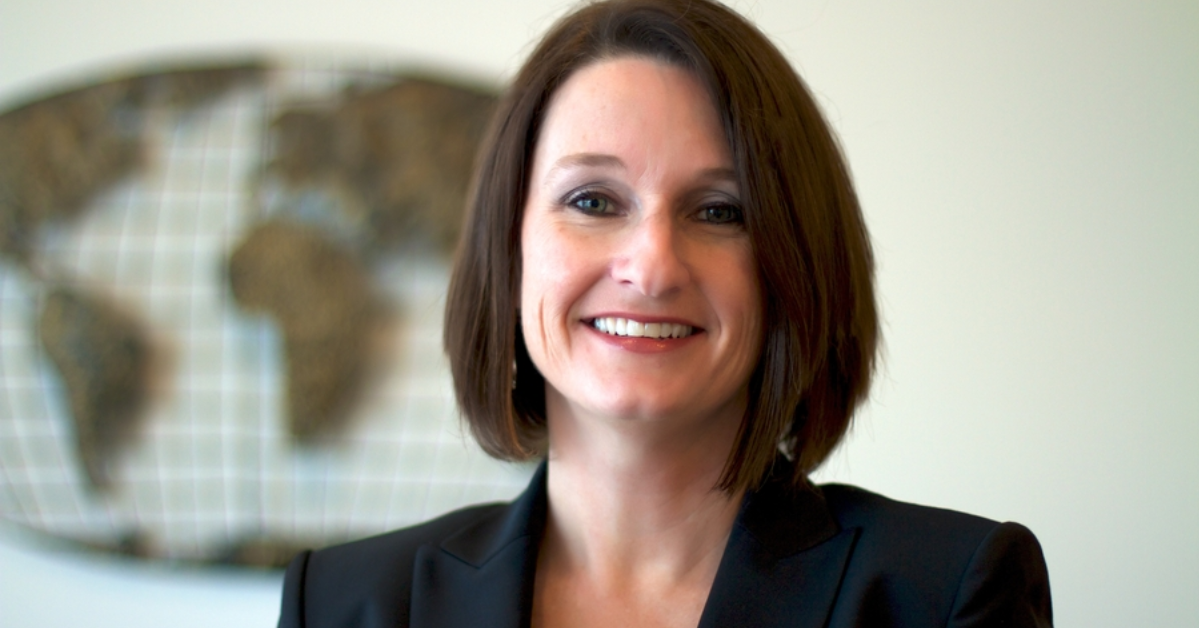 A woman in a black blazer smiles confidently in front of a blurred world map