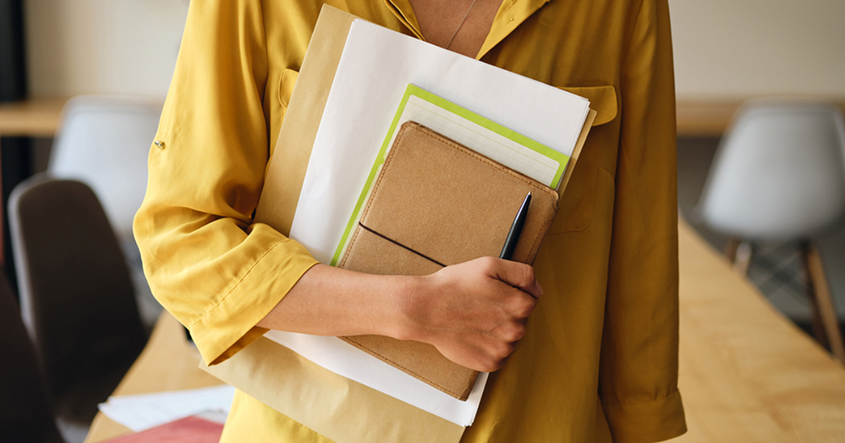 A person in a yellow shirt holds folders and a notebook with a pen