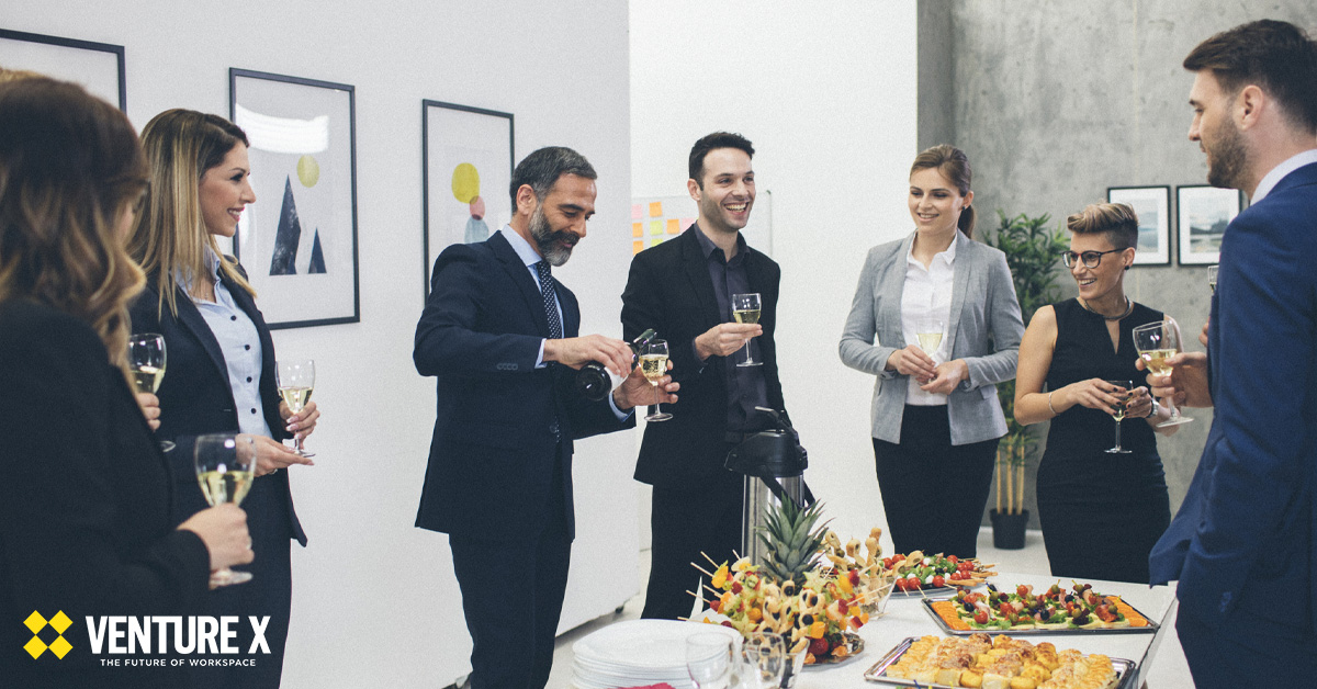 A group of people in business attire stand and hold glasses at a small indoor gathering in a flexible workspace