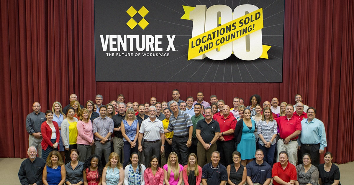 A group of people poses in front of a stage with a large screen displaying "Venture X: The Future of Workspace