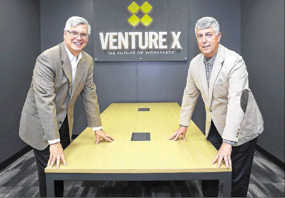 Two men in suits stand on either side of a conference table in front of a Venture X sign