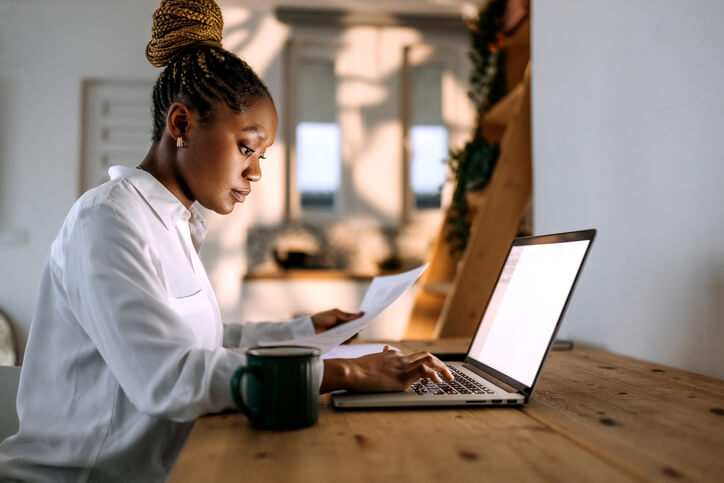 A person with braided hair works on a laptop while holding a document. A green mug is placed on the wooden table beside the laptop. office space for rent near me